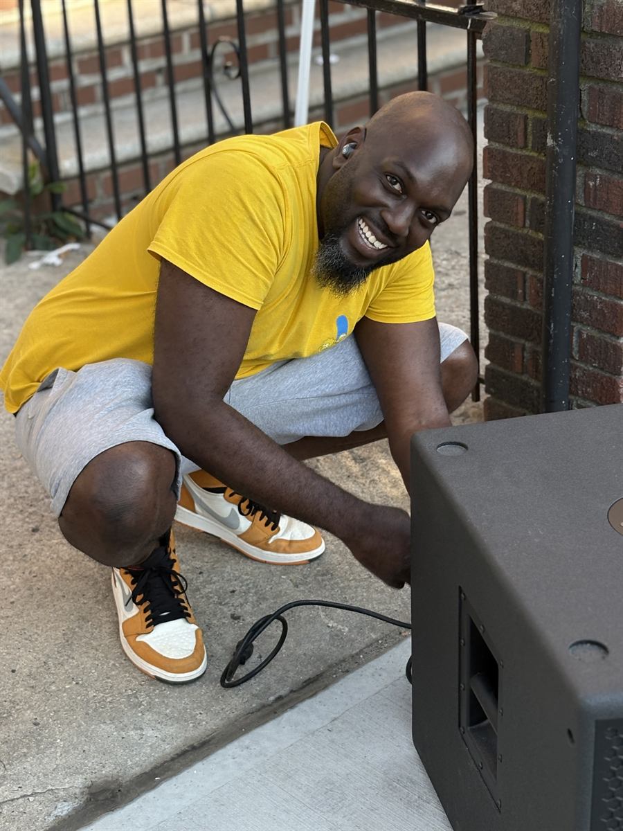 A neighbor in a yellow t-shirt crouched down on the sidewalk hooking up a speaker