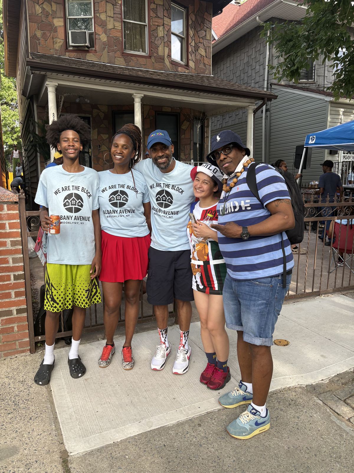 Five neighbors together on the sidewalk in front of a house on the avenue, three of them wearing We Are The Block t-shirts