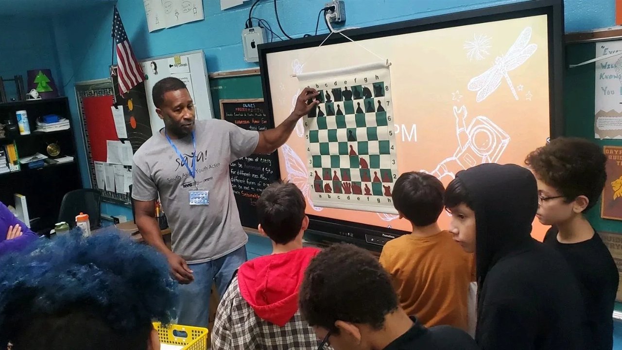 Lamont teaching a chess lesson to a group of kids, working through a position on a demonstration board at the front of the room