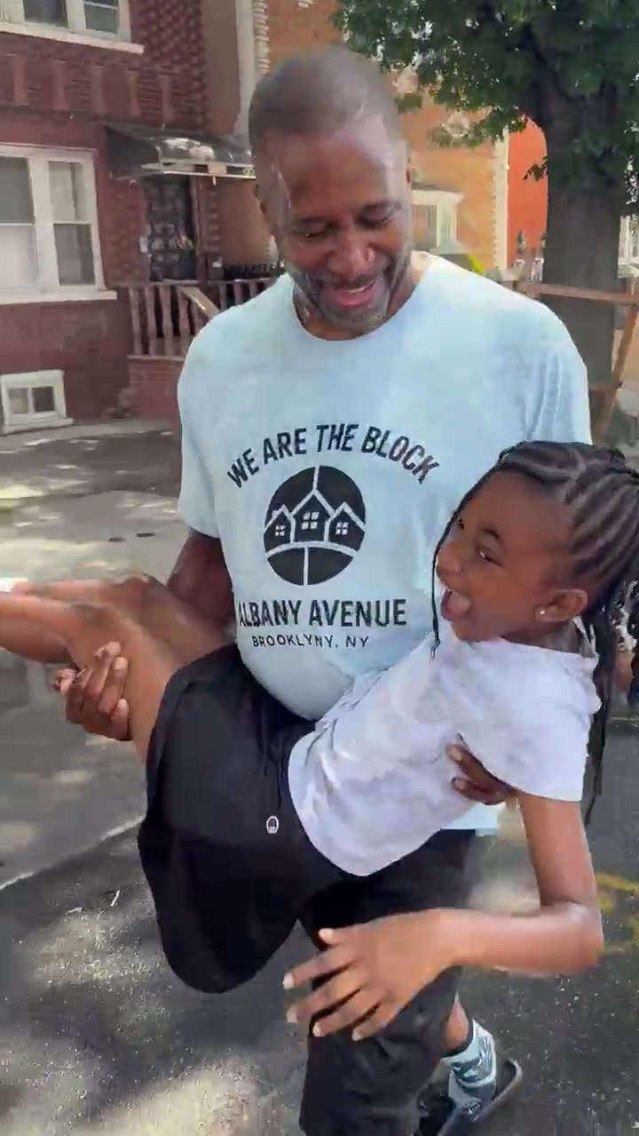 A neighbor in a We Are The Block t-shirt carrying a laughing girl down the middle of the wet street